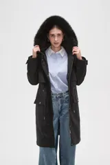 Campera parka negra de corte largo y holgado, con capucha fija adornada con piel sintética, cierre frontal oculto y bolsillos laterales con cordones ajustables en la cintura.