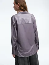 Camisa de corte oversize confeccionada en tejido jersey con acabado brillante. Presenta cuello en punta, cierre frontal con botones, mangas largas con puños anchos y tajos laterales en el ruedo.