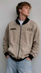 Campera de fleece color beige, con cierre frontal completo, cuello alto, puños, dobladillo y cuello en contraste negro. Cuenta con bolsillos tipo canguro y detalles bordados en el pecho.