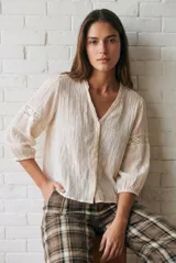 Blusa de color arena con cuello en V, mangas tres cuartos abullonadas y detalles de encaje bordado en hombros y mangas. Presenta un diseño ligero y delicado con cierre frontal.