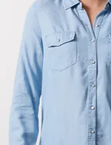 Camisa de manga larga en tela denim de color azul medio, con cuello clásico, botones frontales y dos bolsillos de parche con solapa en el pecho.