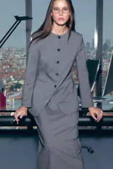 Blazer largo gris oscuro, de corte recto y largo, con escote redondo sin solapa y cierre frontal de botones oscuros. Presenta bolsillos ribeteados a la altura de la cadera y forro interior.