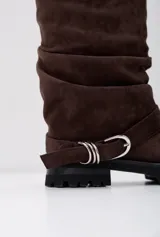Botas tipo borcego de gamuza color marrón oscuro, con diseño de caña media arrugada y suela track negra. Incluye una tira decorativa con hebilla metálica plateada en la zona del tobillo.