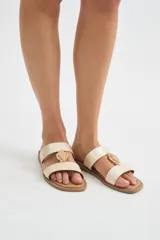 Sandalias planas color crudo con doble tira y detalle de piedra decorativa al centro con borde dorado.