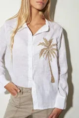 Camisa blanca de lino con manga larga y botones frontales. Presenta un bordado artesanal en hilo color paja que simula una palmera en el lado izquierdo del pecho.