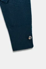 Jersey de punto de tacto suave, color azul petróleo, con cuello subido y manga larga. Presenta detalle de botones metálicos en el hombro y en los puños.