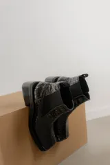 Botas tipo chelsea de cuero color marrón oscuro con acabado brillante. Presentan punta cuadrada con detalle de puntera metálica, elásticos laterales y taco cuadrado bajo.