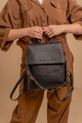 Mochila cartera negra de cuero sintético con textura granulada, tapa frontal con cierre magnético y placa metálica con logo. Cuenta con correas ajustables y una correa de hombro adicional con detalles de ojales metálicos.