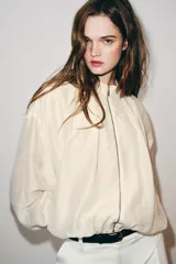 Chaqueta bomber de cuello subido y manga larga, con acabado satinado en color beige. Presenta pliegues frontales, bolsillos laterales ocultos en costura y cierres elásticos en puños y cintura. Cierre frontal con cremallera.