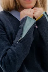 Buzo de lambswool color beige, con escote redondo y detalle en V. Presenta cierres metálicos en los puños que revelan un interior en contraste de color marrón y ribete rojo. Tiene tajos laterales para mayor comodidad y un fit amplio.