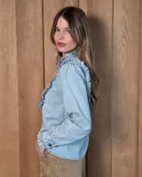 Camisa de denim celeste con cuello alto con volados, botonadura frontal y detalles de volados verticales a lo largo del frente. Cuenta con mangas largas abullonadas y puños con volados.