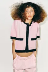 Chaqueta de punto rosa con ribetes negros en el cuello, la botonera, los bolsillos, los puños y el ruedo. Cuenta con manga corta, cuello redondo, bolsillos frontales de plastrón con botones y cierre frontal abotonado.