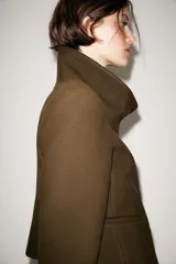 Chaqueta corta de paño color verde militar, con cuello subido, manga larga con puños abotonados y cierre frontal cruzado con botones metálicos. Presenta hombreras y bolsillos de vivo en el delantero.