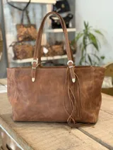Cartera tipo tote con estampado animal print de leopardo. Cuenta con doble asa de hombro ajustable con hebillas metálicas y detalle de flecos colgantes en uno de los laterales.