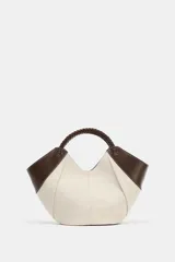 Bolso mini shopper de lona en color crudo con detalles en cuero sintético marrón en los laterales y una manija corta trenzada de mano. Cuenta con costuras contrastantes y cierre magnético.