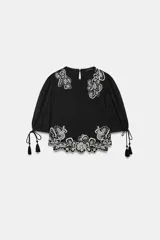Blusa corta negra con mangas abullonadas y bordados florales en contraste de color blanco. Presenta cuello redondo con puntilla, mangas ajustables con cordones y borlas, y cierre en la espalda con abertura y botón oculto.