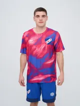 Remera deportiva Umbro del Club Nacional de Football, modelo Press para hombre. Diseño de manga corta con estampado abstracto en tonos rojo, rosado y azul. Incluye el logo de Umbro y el escudo del club en el pecho.