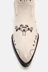 Botín estilo cowboy confeccionado en piel color crudo, con diseño de flores bordadas en tono marrón. Presenta tacón ancho en bloque, acabado en punta y tiradores laterales.