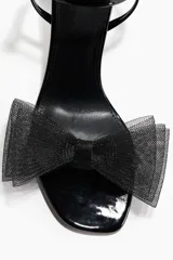 Sandalias de taco alto negro con acabado charolado, tira delantera con detalle de lazo voluminoso de tul y tira ajustable al tobillo con hebilla metálica.