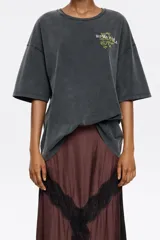 Camiseta de algodón orgánico color gris oscuro con efecto desgastado (acid wash). Presenta un corte oversize, escote redondo y un pequeño estampado gráfico en tono amarillo/verde en el pecho.