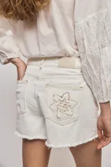Short de denim blanco de calce recto y tiro medio, con bolsillos delanteros, cierre con botón y terminación desflecada en el ruedo. Presenta detalles florales bordados al tono en el frente.