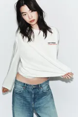 Camiseta de manga larga color blanco, corte regular fit, con cuello redondo y bajo recto. Presenta un pequeño texto estampado en color rojo en el pecho.