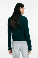 Saco corto de punto trenzado fino en color verde oscuro, con cuello redondo, cierre frontal de botones y bolsillos delanteros. Presenta detalles de ribetes festoneados en color crema en los bolsillos y en los puños.