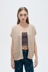 Chaleco de manga corta tejido en acrílico, color beige, con escote en V, cierre frontal de botones y bolsillos delanteros aplicados.
