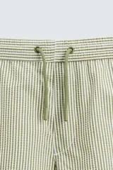Bañador de hombre color verde claro con finas rayas blancas verticales (efecto seersucker). Tiene cintura elástica ajustable con cordón verde, bolsillos laterales y un bolsillo trasero de vivo.