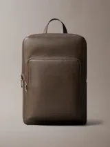 Mochila de hombre con silueta slim y cuadrada, confeccionada en material sintético con textura microgranulada (micro pebble). Color marrón oscuro, con asa de mano superior, correas acolchadas para el hombro, cierre de cremallera doble y bolsillo exterior frontal con cierre.
