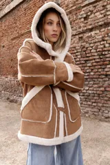 Campera estilo gamulán color tostado con interior y bordes de corderito sintético color crema. Presenta capucha amplia, cierre frontal con trabas y broches metálicos, y bolsillos delanteros grandes. Posee un calce relajado y diseño de textura suave.