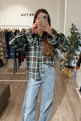 Camisa de manga larga a cuadros en tonos verde, negro y blanco, con diseño de corte holgado, cuello clásico, cierre frontal con botones y detalle de volados en los hombros con puños elastizados.