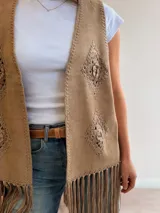 Chaleco de gamuza sintética color marrón, sin mangas, con diseño abierto, bordados geométricos de crochet en los laterales y largos flecos en el ruedo.