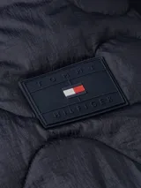 Campera acolchada azul marino de nailon con aislamiento térmico, repelente al agua, capucha fija con cordón ajustable, cierre de cremallera, dos bolsillos diagonales en la cintura y parche con el logo de Tommy Hilfiger en la manga.