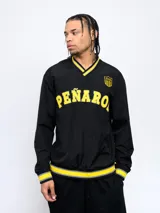 Buzo negro de nylon con cuello en V, puños y pretina con detalles en amarillo y gris. Estampado frontal con la palabra "Peñarol" en amarillo y escudo del club en la manga. Corte clásico.