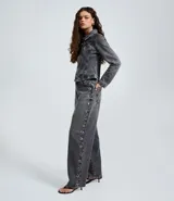 Pantalón baggy de jean color gris desteñido, con corte amplio y recto. Presenta una hilera de ojales metálicos decorativos a lo largo de los laterales y cierre frontal con botón y cremallera.