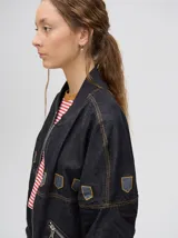 Campera tipo bomber confeccionada en denim de algodón color negro. Presenta un diseño holgado con cierre frontal metálico, bolsillos laterales con cierre y detalles decorativos en forma de escudos en la parte superior delantera.