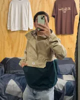 Campera rompeviento de diseño bicolor, con parte superior en tono arena y parte inferior en verde oscuro. Cuenta con capucha, cuello alto, cierre frontal parcial oculto y un amplio bolsillo frontal tipo canguro.