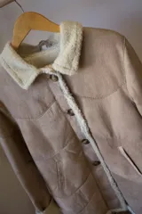 Campera tipo gamulán de cuero genuino en color marrón, con interior y detalles en corderito sintético color crema. Presenta cierre frontal con botones, cuello solapa y puños con vuelta.