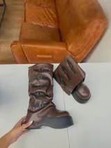 Botas bucaneras de cuero vegano color marrón, con caña arrugada y plataforma gruesa negra. Presentan cinco tiras ajustables con hebillas doradas a lo largo de la caña.