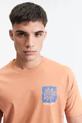 Remera de manga corta color salmón, con escote redondo y un pequeño estampado frontal de una flor azul sobre un fondo texturizado, con la inscripción "MISTRAL ® 1976 INTERNATIONAL".