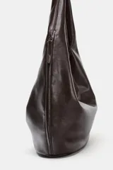 Bolso de hombro tipo saca en formato maxi, confeccionado en material sintético negro. Presenta un diseño holgado con asa integrada, cierre imantado en el compartimento principal, bolsillo lateral con cremallera y detalle de tirador.