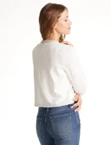 Chaqueta tejida blanca de la marca Rio & Rian, con cuello a la base bordado con perlas y canutillos. Cuenta con broche de enganche interno, bolsillos simulados en la delantera con aplique de brillos, y vivo delantero y puños en tejido lurex.