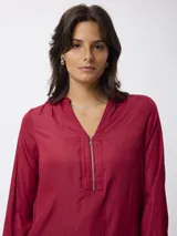 Blusa de satén color azul, con cuello camisero y cierre frontal parcial con cremallera. Presenta mangas largas con puños abotonados y un corte recto y fluido.