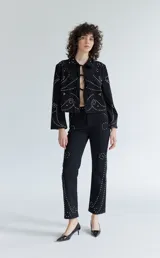 Pantalón de denim negro con diseño de tachas metálicas distribuidas en patrones curvos. Presenta un corte recto y tiro medio.