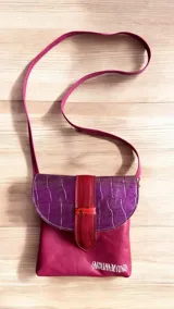 Bandolera de cuero natural de diseño compacto y formato rectangular con solapa. Presenta cuerpo fucsia, solapa superior de cuero grabado con textura simil cocodrilo en tonos morados, y tira central vertical en tono bordó con detalle pintado a mano. Incluye correa larga cruzada ajustable y el logo de la marca estampado en blanco en el frente inferior.