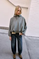 Chaqueta estilo bomber de corte oversize en color marrón oscuro, confeccionada en tejido suave. Presenta capucha con cordones ajustables, cierre frontal con cremallera, mangas largas con puños elásticos y bolsillos delanteros de vivo. El bajo cuenta con detalle de pliegues.