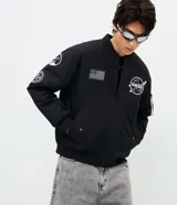 Campera bomber negra de microfibra, con cierre frontal por cremallera, puños y ruedo elásticos, y bolsillos con cierre. Cuenta con múltiples parches y estampas bordadas alusivas a la NASA, incluyendo el logo principal en el pecho, insignias en la manga derecha y una bandera de Estados Unidos.