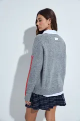 Sweater gris de calce relajado con cuello redondo y mangas largas. Presenta detalles de rayas horizontales en color rojo a lo largo de las mangas, con puños y ruedo acanalados.