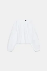 Camisa blanca de popelín, manga larga abullonada con puño ajustado y cuello redondo. Presenta frunces en el canesú delantero y trasero, creando un efecto holgado y corto.